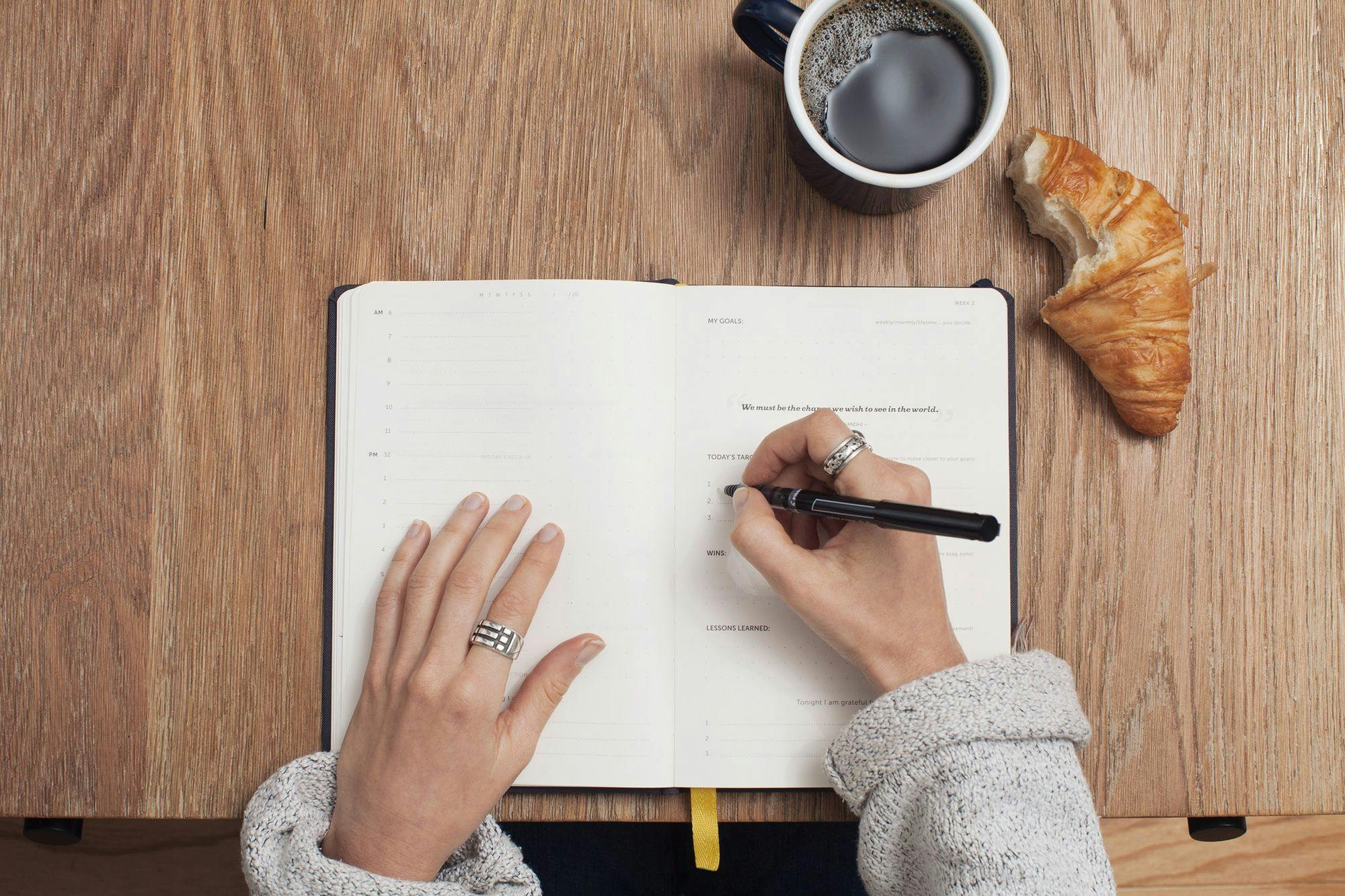 woman-writing-in-notepad-list-with-black-pen-coffee-croissant-and-rings-on-fingers